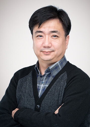 A man with short black hair is smiling at the camera, wearing a gray sweater over a blue and white plaid shirt. (Captioned by AI)