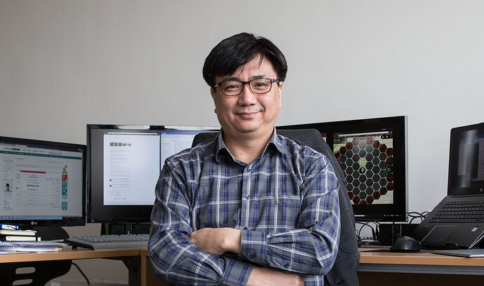 A man with glasses is seated at an office desk, arms crossed, with monitors displaying a website and a patterned design. (Captioned by AI)