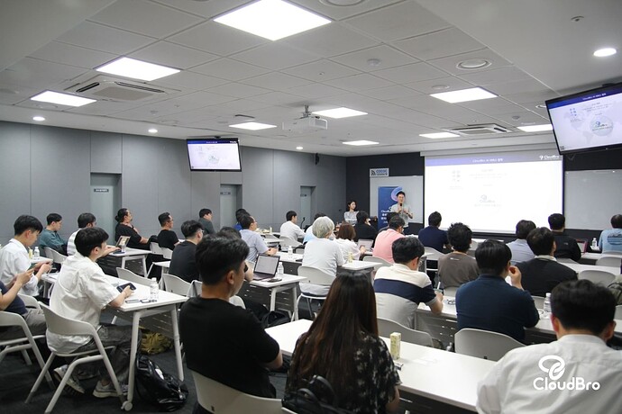 The image shows a classroom or meeting room filled with people attentively listening to a speaker who is giving a presentation on a stage with a projection screen displaying information, in a modern, well-lit indoor setting. (Captioned by AI)