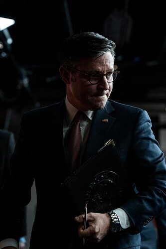Mike Johnson holds a folder after a press conference. He’s wearing a dark blue suit with a maroon tie.